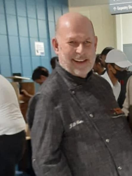 John-David Skene smiling, wearing a charcoal chef’s jacket embroidered with his initials.