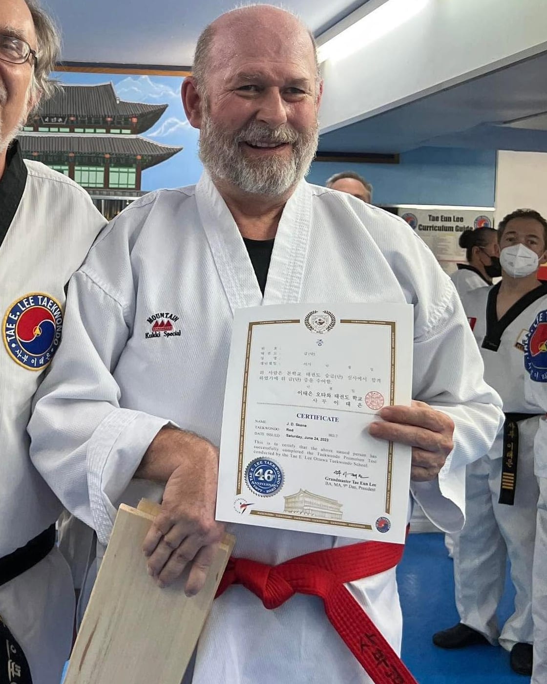 John-David Skene in his Taekwondo uniform and red belt, smiling as he holds a promotion certificate and a broken wooden board.